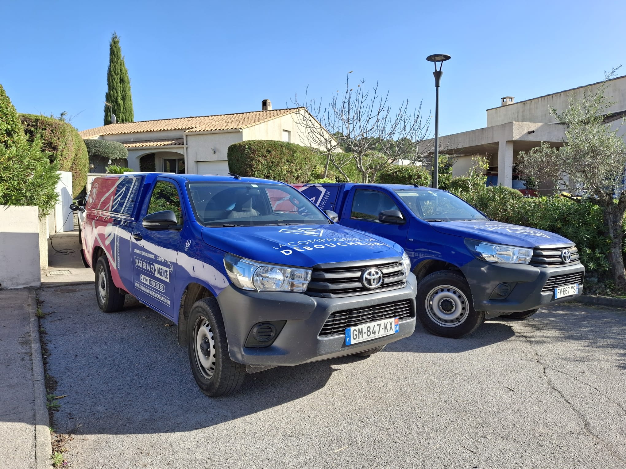 Deux pick-up bleus de La Compagnie Déboucheurs stationnés devant une maison à Montpellier.
