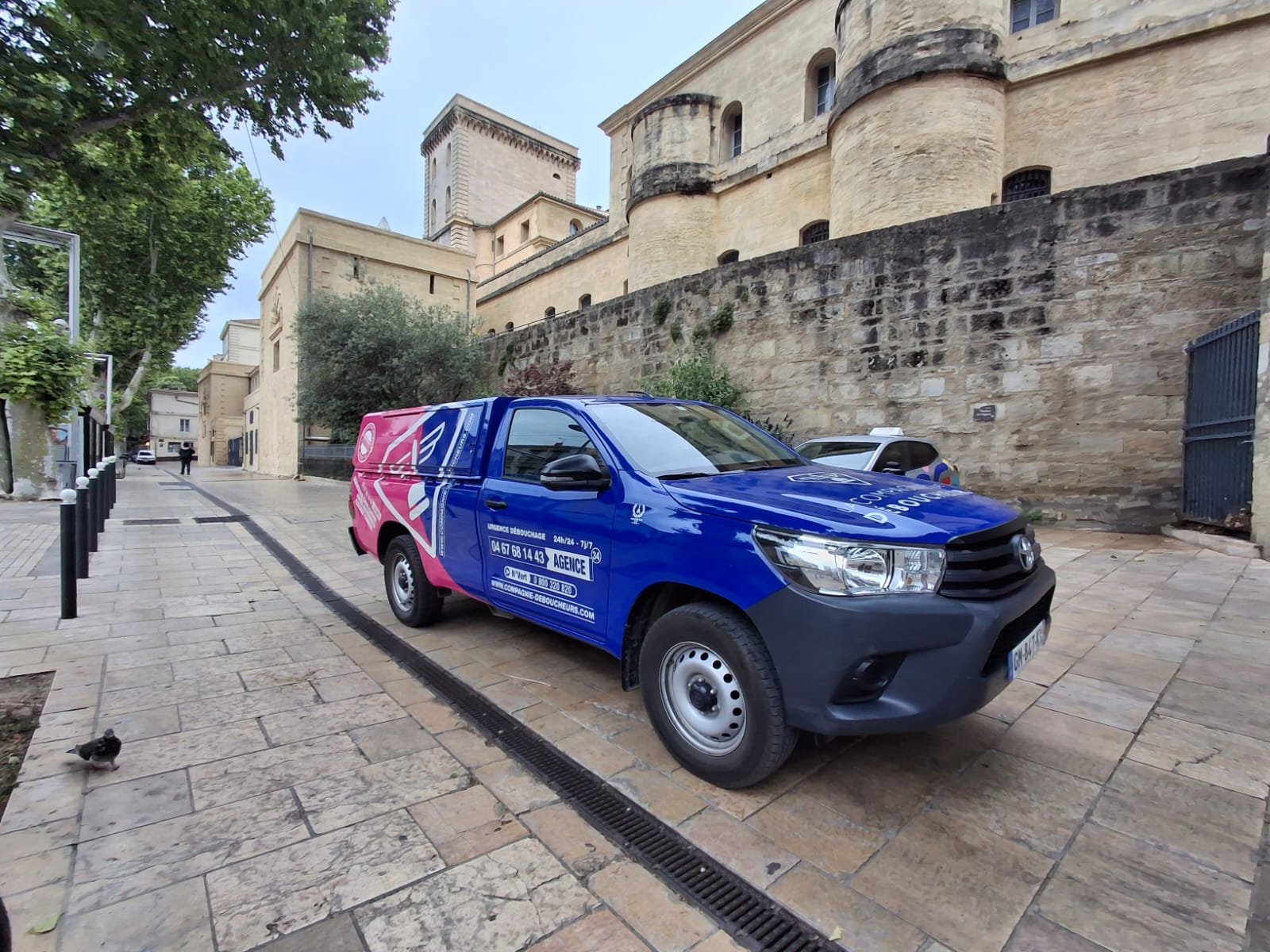 Pick-up bleu et rose de La Compagnie Déboucheurs stationné dans une rue pavée à Montpellier, devant un bâtiment historique.