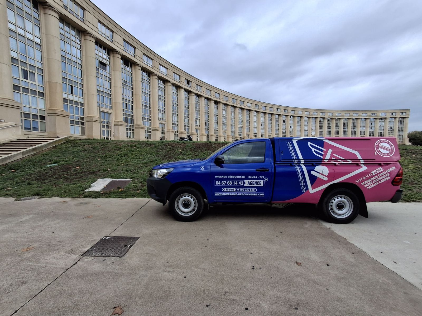 Pick-up bleu et rose de La Compagnie Déboucheurs stationné devant un grand bâtiment institutionnel à Montpellier.